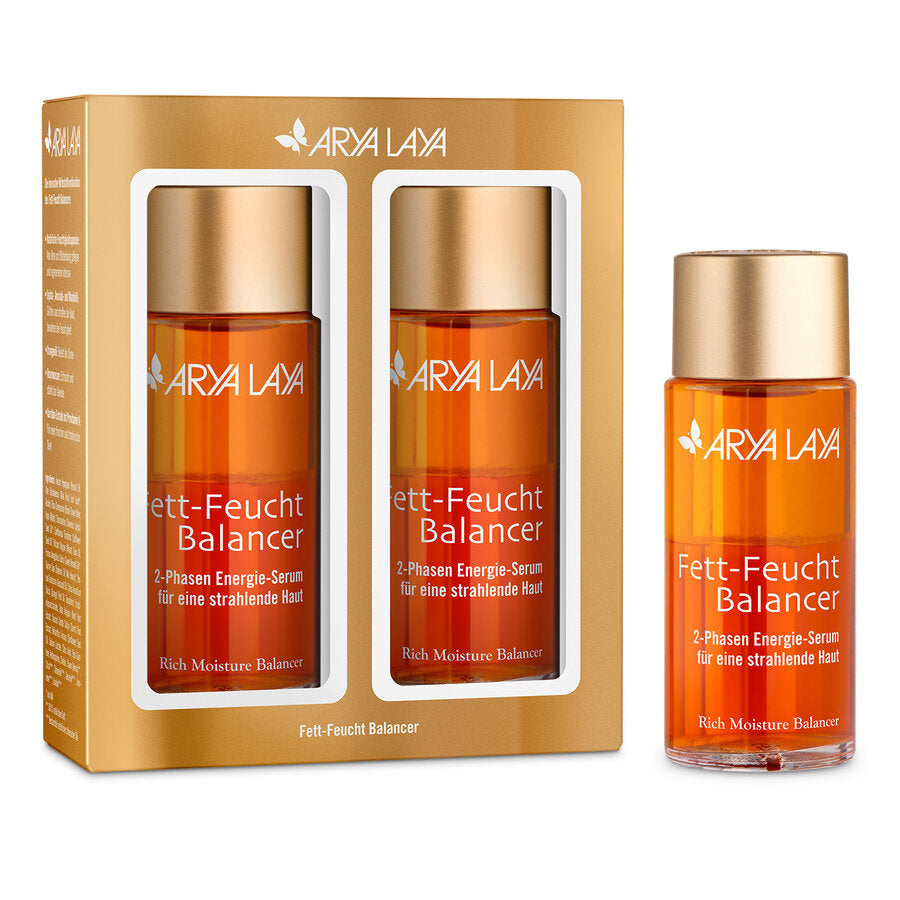 Eine Doppelpackung des Arya Laya Fett-Feucht Balancer 2-Phasen Energie-Serums. Die Verpackung ist goldfarben und enthält zwei transparente Flaschen mit goldenem Schraubverschluss. Das Etikett trägt die Aufschrift „Fett-Feucht Balancer – 2-Phasen Energie-Serum für eine strahlende Haut“ und „Rich Moisture Balancer“. Neben der Verpackung steht eine einzelne Flasche des Serums.