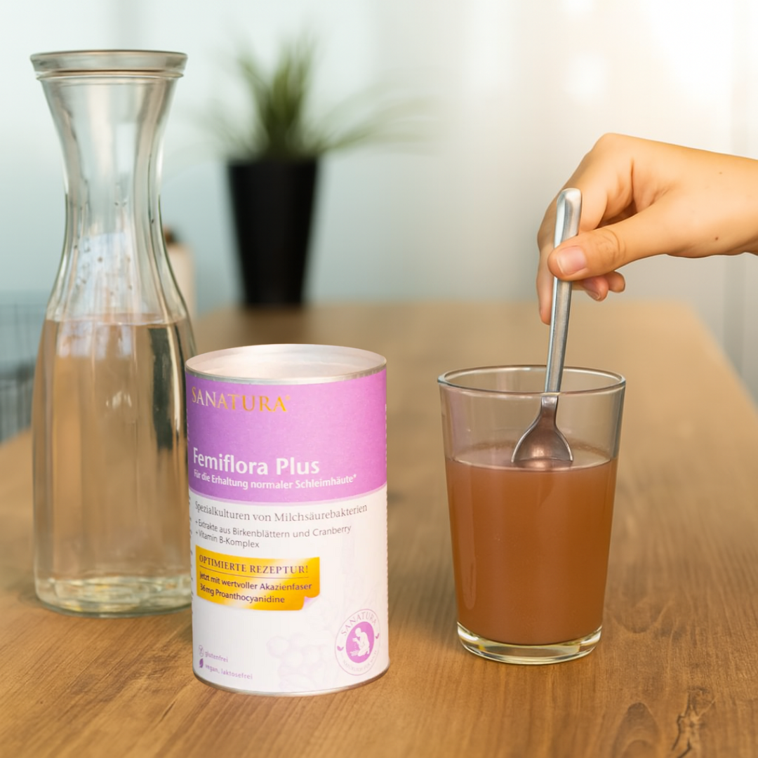 Sanatura Femiflora Plus in der Anwendung – Nahrungsergänzungspulver wird in einem Glas mit Wasser angerührt, daneben Dose und Karaffe auf Holzoberfläche