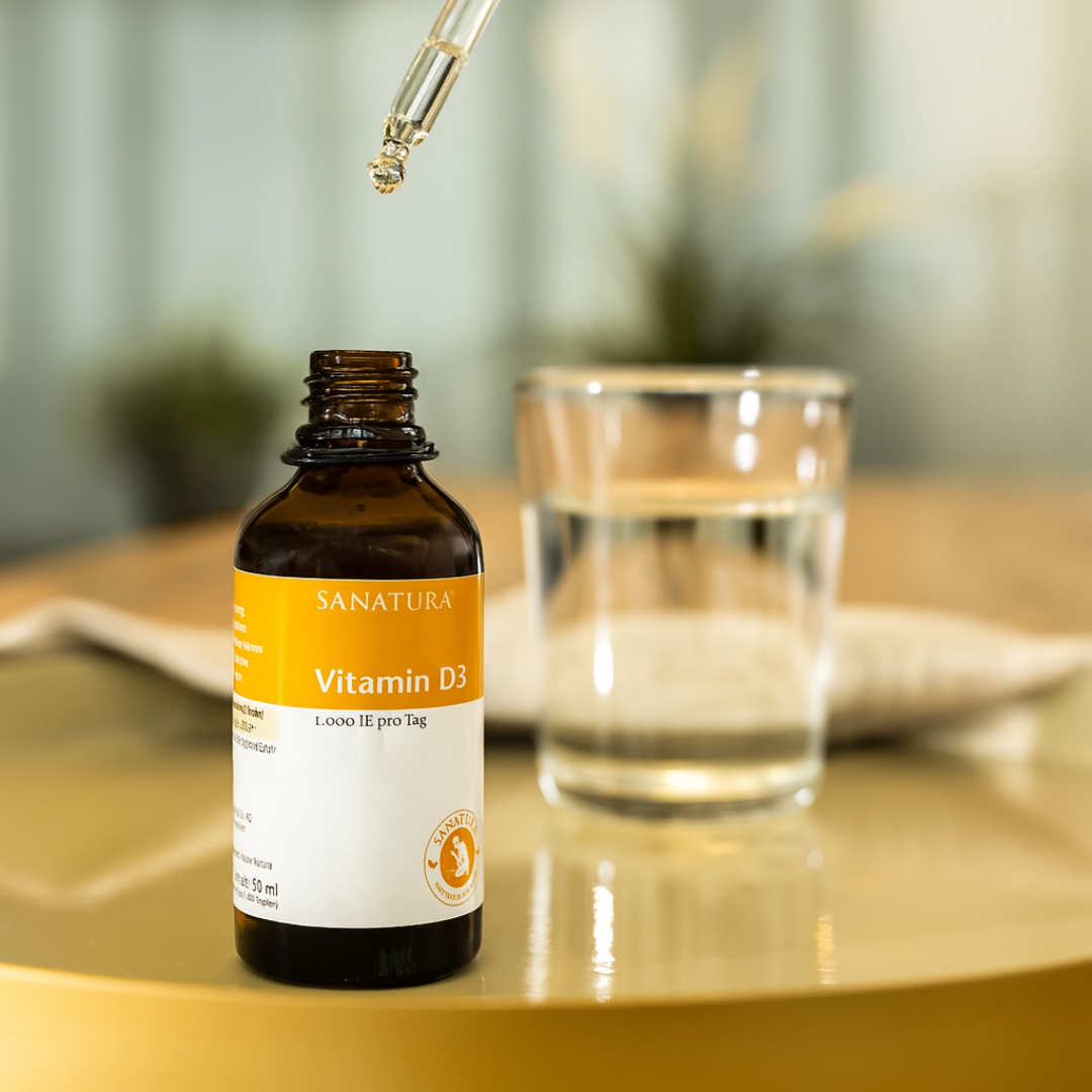 Anwendung der Sanatura Vitamin D3 Tropfen mit Pipette – eine Tagesdosis in einem Glas Wasser auf einem Tisch mit natürlichem Hintergrund