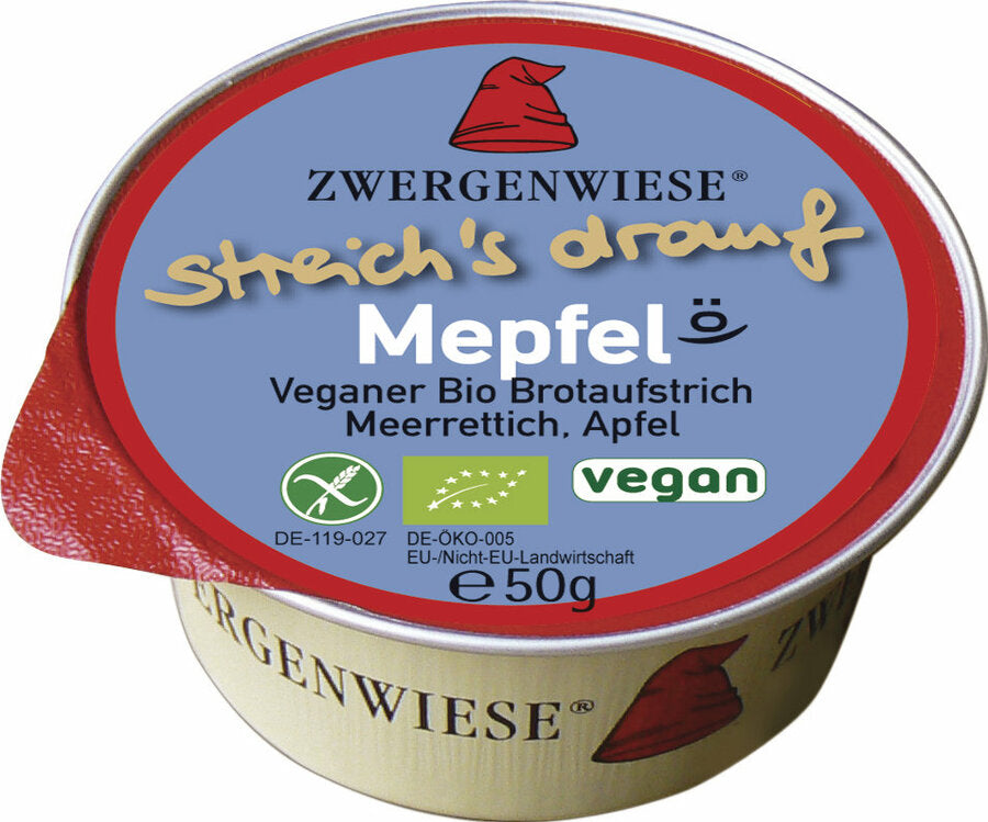 Das Bild zeigt eine runde, vegane Brotaufstrich-Dose der Marke Zwergenwiese. Die Dose hat einen roten Deckelrand und eine blaue Oberseite mit der Aufschrift "streich's drauf Mepfel". Unterhalb des Produktnamens steht "Veganer Bio Brotaufstrich Meerrettich, Apfel". Es gibt auch Symbole, die das Produkt als vegan und bio-zertifiziert kennzeichnen. Die Nettofüllmenge beträgt 50 Gramm. Das Markensymbol, ein roter Zwergenhut, ist ebenfalls auf dem Deckel abgebildet.