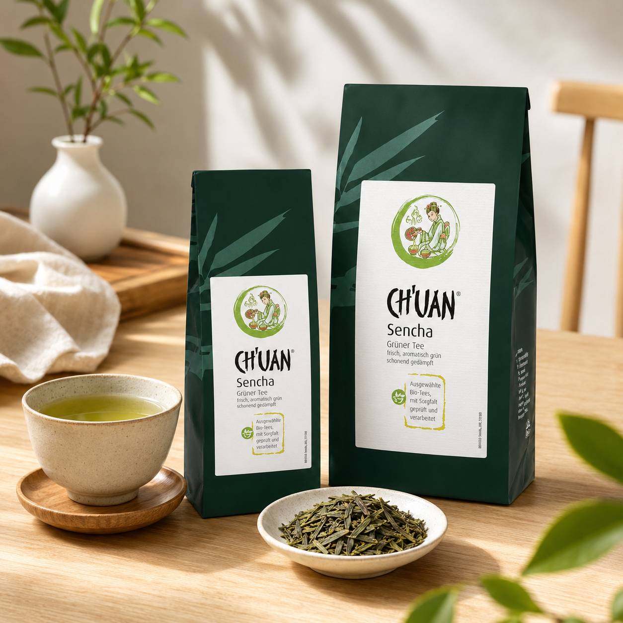 CH'UAN Sencha Bio Grüntee in zwei Größen mit Teeschale und losen Teeblättern auf einem Holztisch