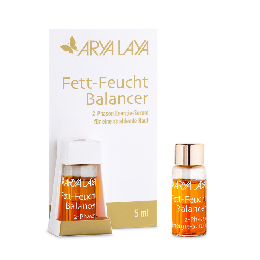 Arya Laya Fett-Feucht Balancer 2-Phasen Energie-Serum in einer 5-ml-Miniaturflasche mit goldfarbenem Deckel. Die Verpackung ist weiß mit goldener Schrift. Eine der kleinen Flaschen ist in einer weißen Halterung befestigt, daneben steht eine weitere Mini-Flasche mit orangefarbenem Serum.