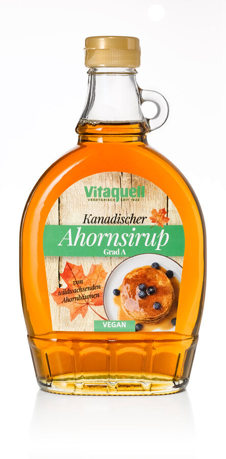 Eine Glasflasche Vitaquell Kanadischer Ahornsirup Grad A mit goldenem Schraubverschluss und Henkel. Das Etikett zeigt eine Holzoptik mit einem Ahornblatt und einem Teller mit Pancakes und Blaubeeren. Der Sirup wird als „vegan“ und aus „wildwachsenden Ahornbäumen“ stammend beschrieben.