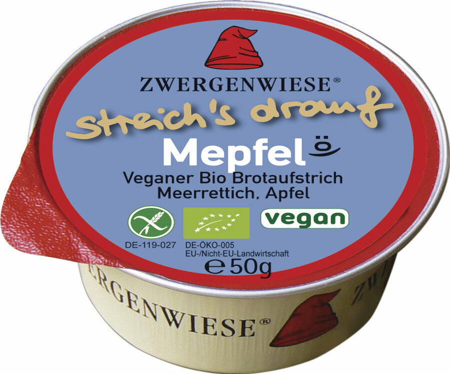  Das Bild zeigt eine runde, vegane Brotaufstrich-Dose der Marke Zwergenwiese. Die Dose hat einen roten Deckelrand und eine blaue Oberseite mit der Aufschrift "streich's drauf Mepfel". Unterhalb des Produktnamens steht "Veganer Bio Brotaufstrich Meerrettich, Apfel". Es gibt auch Symbole, die das Produkt als vegan und bio-zertifiziert kennzeichnen. Die Nettofüllmenge beträgt 50 Gramm. Das Markensymbol, ein roter Zwergenhut, ist ebenfalls auf dem Deckel abgebildet.