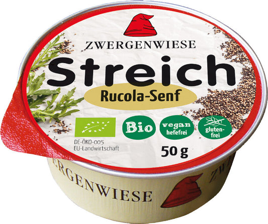 Das Bild zeigt eine runde Dose des veganen Brotaufstrichs "Streich Rucola-Senf" von Zwergenwiese. Die Dose hat einen roten Deckelrand und eine weiße Oberseite mit der Aufschrift "Zwergenwiese Streich Rucola-Senf". Es gibt Symbole, die das Produkt als bio, vegan (hefefrei) und glutenfrei kennzeichnen. Die Nettofüllmenge beträgt 50 Gramm. Auf dem Deckel sind außerdem Abbildungen von Rucola und Senfkörnern zu sehen. Das Markensymbol, ein roter Zwergenhut, ist ebenfalls auf dem Deckel abgebildet.