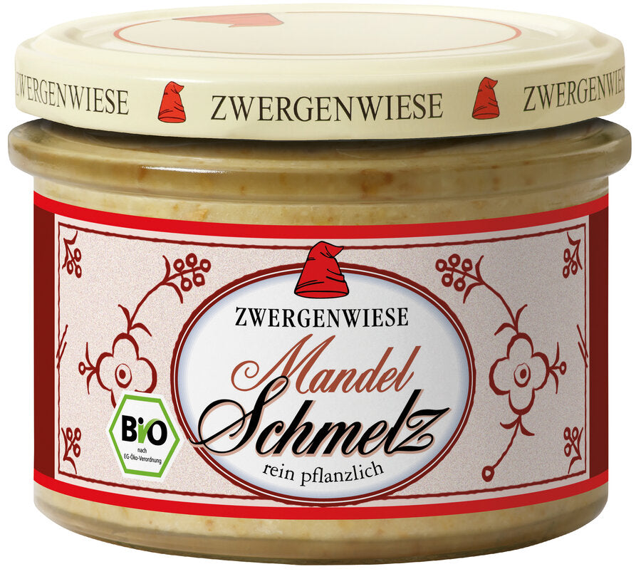 Das Bild zeigt ein Glas "Mandel Schmelz" von Zwergenwiese. Der Deckel ist beige mit einem roten Zwergenhut. Das Etikett ist weiß mit roten Verzierungen, der Aufschrift "ZWERGENWIESE Mandel Schmelz" und einem Bio-Siegel. Darunter steht "rein pflanzlich".