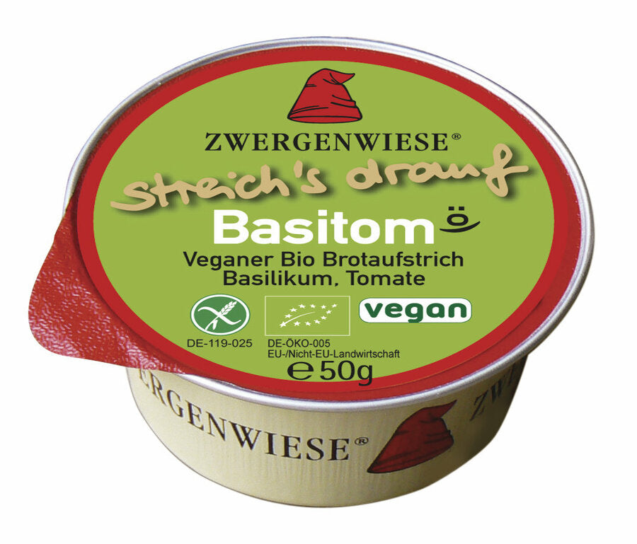 Das Bild zeigt eine runde, vegane Brotaufstrich-Dose der Marke Zwergenwiese. Die Dose hat einen roten Deckelrand und eine grüne Oberseite mit der Aufschrift "streich's drauf Basitom". Unterhalb des Produktnamens steht "Veganer Bio Brotaufstrich Basilikum, Tomate". Es gibt auch Symbole, die das Produkt als vegan und bio-zertifiziert kennzeichnen. Die Nettofüllmenge beträgt 50 Gramm. Das Markensymbol, ein roter Zwergenhut, ist ebenfalls auf dem Deckel abgebildet.