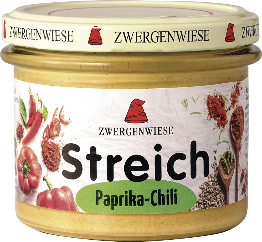 Das Bild zeigt ein Glas des veganen Brotaufstrichs "Streich Paprika-Chili" von Zwergenwiese. Der Deckel ist beige mit einem roten Zwergenhut. Das Etikett ist weiß mit Abbildungen von Paprika, Chili und Gewürzen. In der Mitte steht "Zwergenwiese Streich Paprika-Chili" in schwarzer und grüner Schrift.