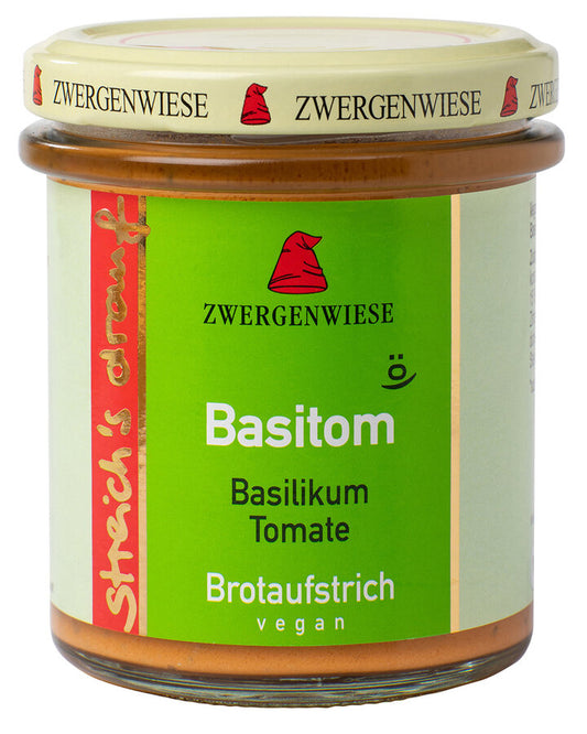 Das Bild zeigt ein Glas "Basitom" Brotaufstrich von Zwergenwiese. Der Deckel ist beige mit einem roten Zwergenhut. Das Etikett ist grün und zeigt die Aufschrift "Zwergenwiese Basitom" in weißer und schwarzer Schrift. Darunter steht "Basilikum Tomate". An der Seite des Etiketts steht "Streich's drauf" in gelber Schrift auf rotem Hintergrund. Der Brotaufstrich ist vegan.