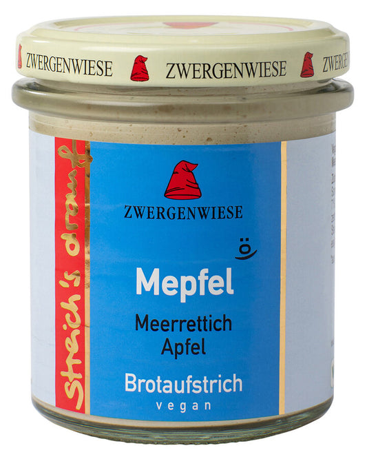 Das Bild zeigt ein Glas "Mepfel" Brotaufstrich von Zwergenwiese. Der Deckel ist beige mit einem roten Zwergenhut. Das Etikett ist blau und zeigt die Aufschrift "Zwergenwiese Mepfel" in weißer Schrift. Darunter steht "Meerrettich Apfel". An der Seite des Etiketts steht "Streich's drauf" in gelber Schrift auf rotem Hintergrund. Der Brotaufstrich ist vegan.