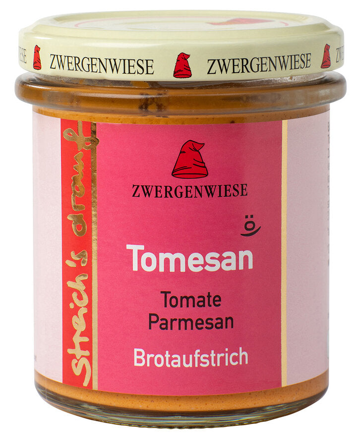 Das Bild zeigt ein Glas "Tomesan" Brotaufstrich von Zwergenwiese. Der Deckel ist beige mit roten Zwergenhüten. Das Etikett ist rosa und zeigt die Aufschrift "Zwergenwiese Tomesan" in weißer Schrift. Darunter steht "Tomate Parmesan". An der Seite des Etiketts steht "Streich's drauf" in gelber Schrift auf rotem Hintergrund.