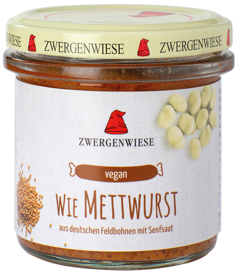  Ein Glas mit dem veganen Aufstrich "Wie Mettwurst" von der Marke Zwergenwiese. Das Etikett zeigt eine rote Zwergenmütze und die Aufschrift "Wie Mettwurst - aus deutschen Feldbohnen mit Senfsaat". Abgebildet sind Feldbohnen und Senfkörner, die die Zutaten und den würzigen Geschmack des Aufstrichs hervorheben. Der beige Deckel trägt mehrfach das Zwergenwiese-Logo. 