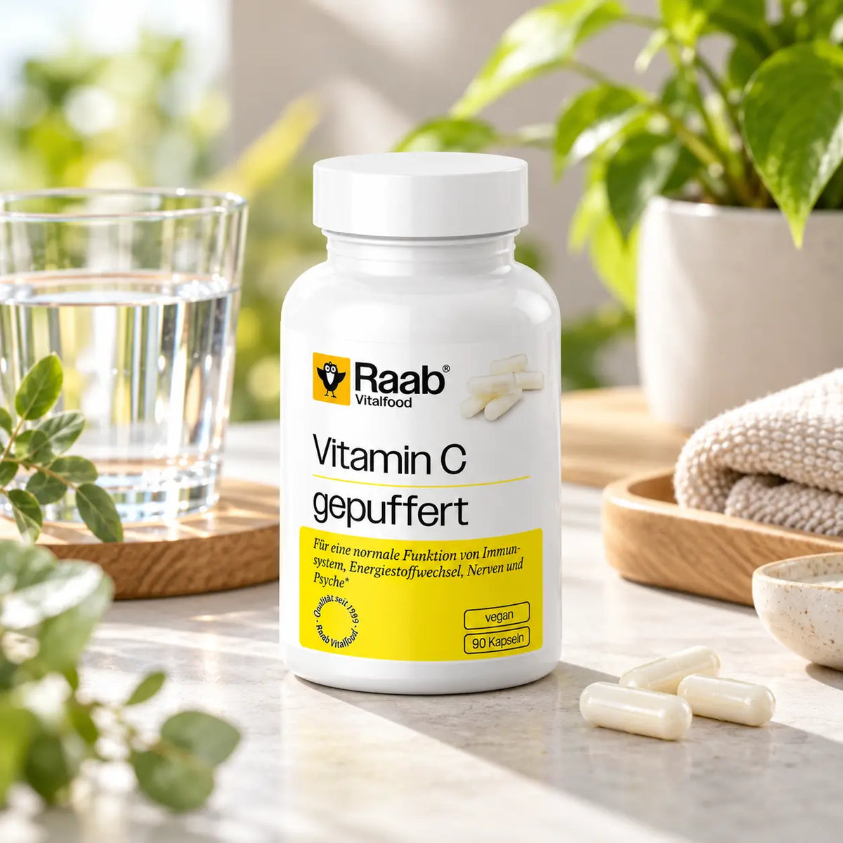 Raab Vitalfood Vitamin C gepuffert in einer hellen Wellness-Umgebung mit Wasserglas, veganen Kapseln und grünen Pflanzen als Teil einer täglichen Nahrungsergänzungsroutine.