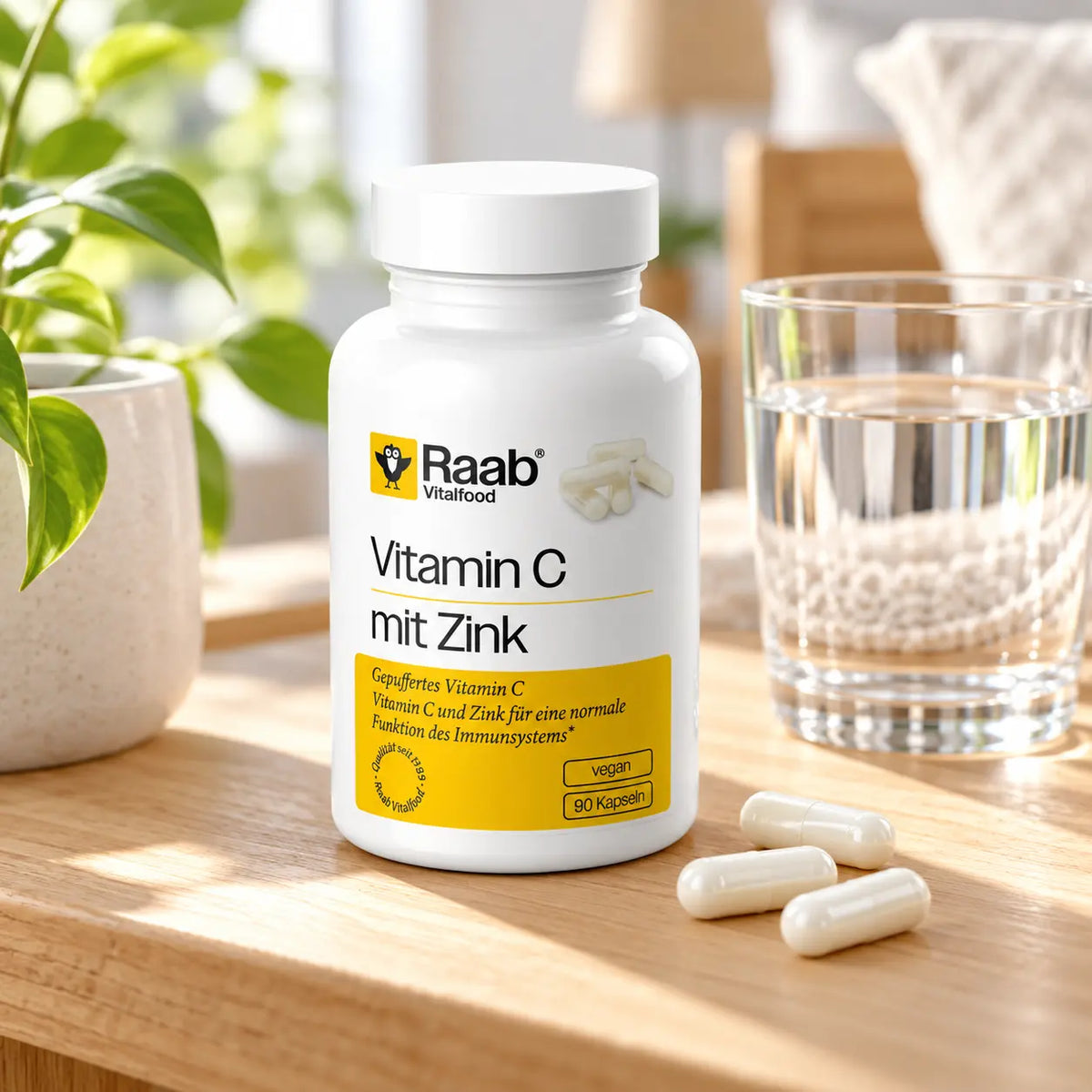 Raab Vitamin C mit Zink, 90 Kapseln auf einem Holztisch mit Wasserglas und einzelnen Kapseln. Nahrungsergänzungsmittel für die tägliche Einnahme.
