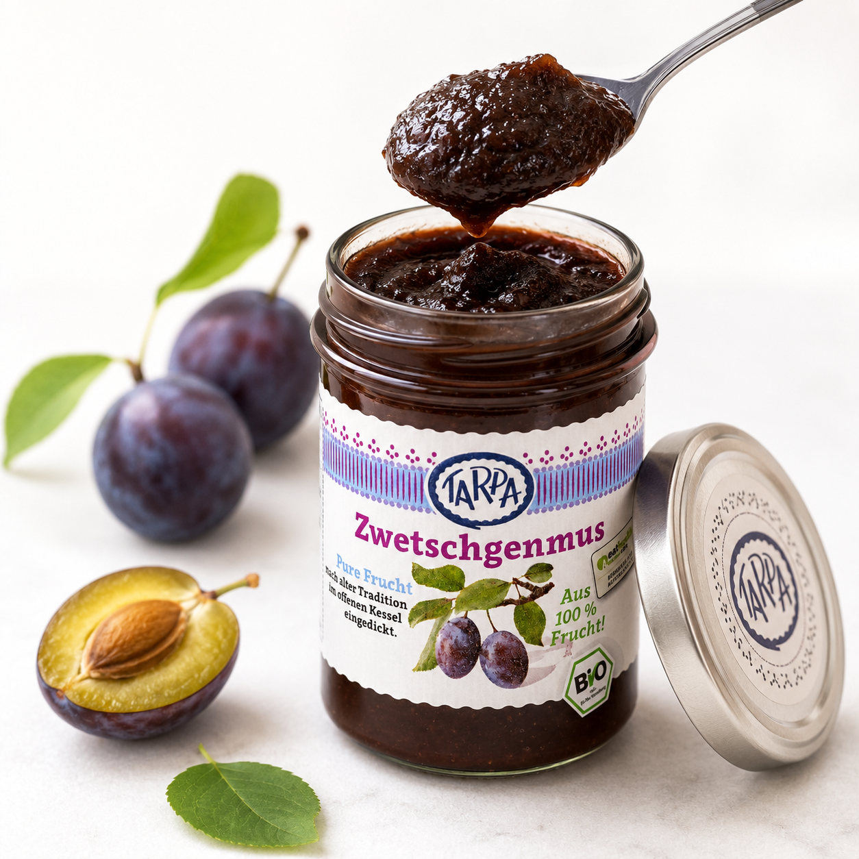 Geöffnetes Glas Tarpa Bio Zwetschgenmus mit Löffel und reifen Zwetschgen